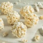 Several classic popcorn balls rest on a sheet of parchment paper atop a wooden surface, with a white spatula nearby and some scattered loose popcorn pieces—perfect for an after-school snack.