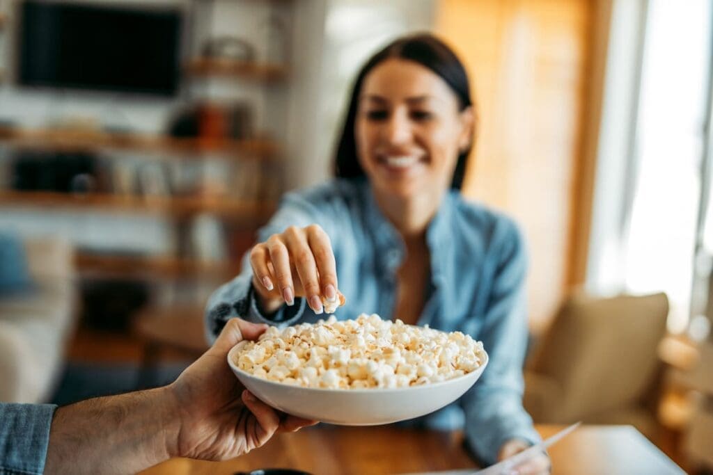 A woman smiles and reaches for popcorn from a bowl held by another person, sitting indoors in a cozy, well-lit living room, while they discuss if popcorn is gluten free.