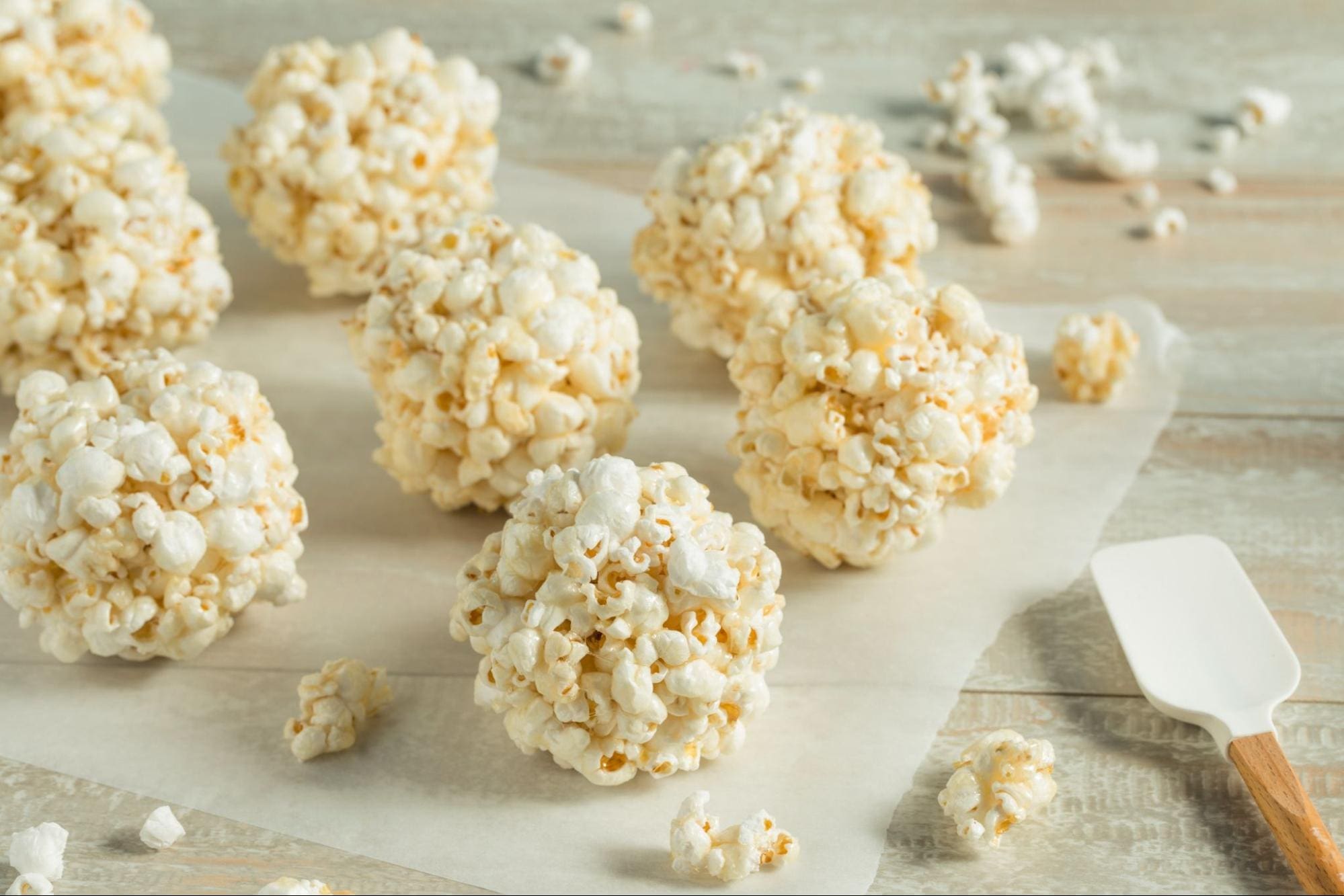 Several classic popcorn balls rest on a sheet of parchment paper atop a wooden surface, with a white spatula nearby and some scattered loose popcorn pieces—perfect for an after-school snack.