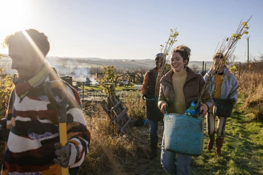 A group of people walk outdoors in sunlight, carrying young trees and gardening tools, smiling and dressed warmly. Partnering for Change, they traverse a rural landscape with hills, smoke, and gardening plots visible in the background.