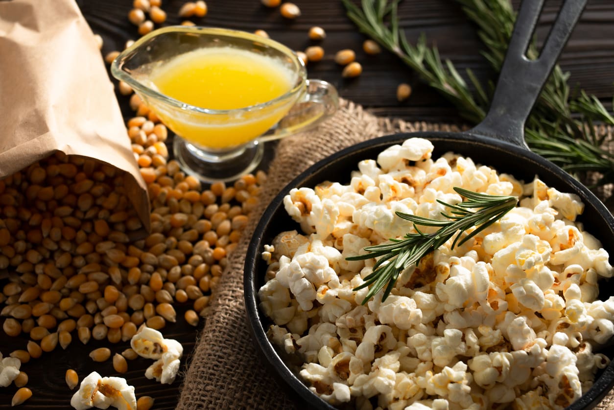 A black skillet filled with gluten free popcorn garnished with rosemary, next to a brown paper bag spilling corn kernels and a small glass container of melted butter, all arranged on a rustic surface.