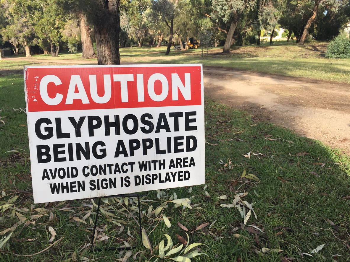 A warning sign on grass reads, CAUTION: Glyphosate being applied. Avoid contact with area when sign is displayed. Trees and a dirt path are visible in the background, highlighting concerns about glyphosates possible effects on gut health.