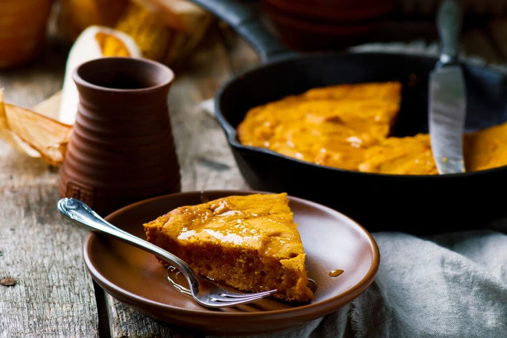 A slice of honey skillet cake drizzled with honey sits on a brown plate with a fork, next to a rustic mug and a cast iron skillet containing the rest of the cornmeal skillet cake on a wooden table.