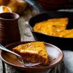 A slice of honey skillet cake drizzled with honey sits on a brown plate with a fork, next to a rustic mug and a cast iron skillet containing the rest of the cornmeal skillet cake on a wooden table.