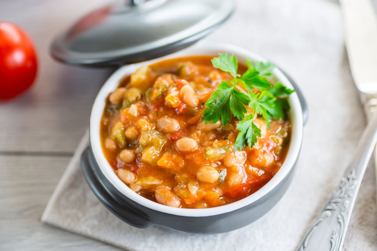 A bowl of vegetable and bean stew garnished with fresh parsley, served in a black dish with a lid beside it, resting on a folded napkin with a silver knife nearby.