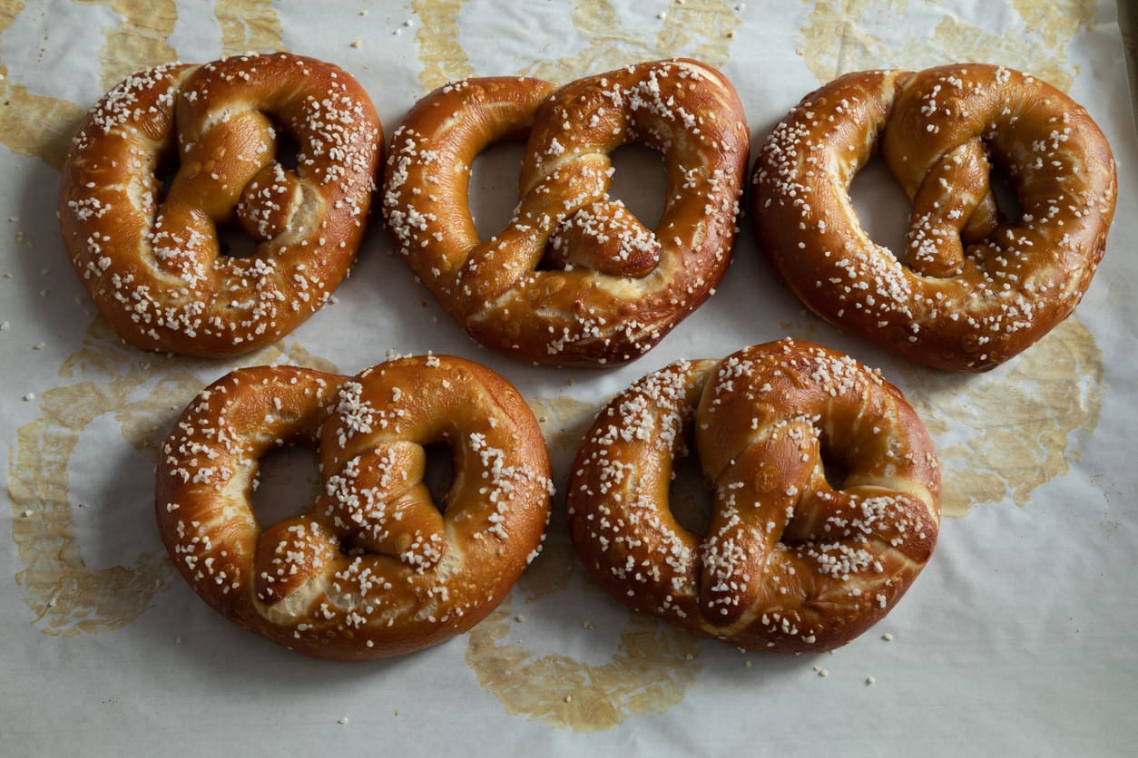 Five golden-brown soft pretzels topped with coarse salt are arranged on a baking sheet lined with parchment paper, perfect for Oktoberfest celebrations, with visible grease marks surrounding them.