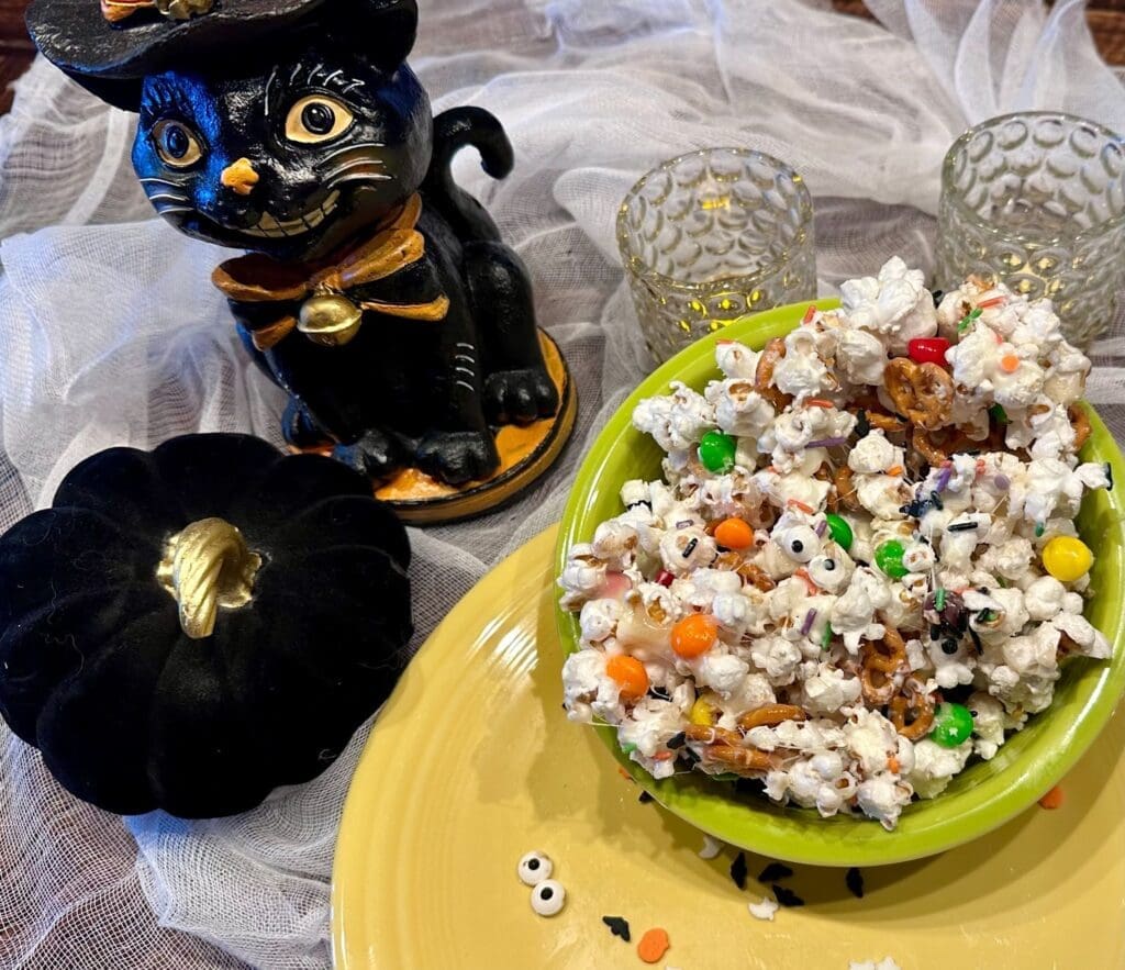 A bowl of Halloween snack mix with organic popcorn, candy, and pretzels sits on a yellow plate next to a black cat figurine, a black pumpkin, and two glass candle holders on a white mesh cloth.
