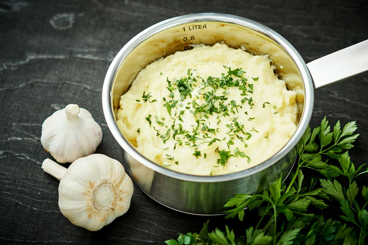 A pot of creamy mashed potatoes garnished with chopped parsley sits on a dark surface, accompanied by garlic bulbs and fresh parsley—perfect for Thanksgiving sides.
