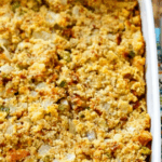 A close-up of a southern style cornbread stuffing in a white casserole dish, showing a golden-brown, crumbly texture with visible pieces of onion and celery.