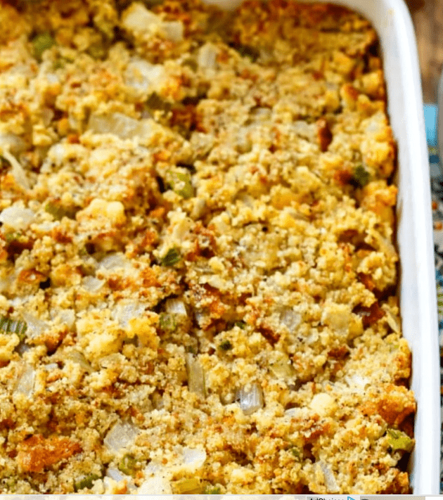 A close-up of a southern style cornbread stuffing in a white casserole dish, showing a golden-brown, crumbly texture with visible pieces of onion and celery.