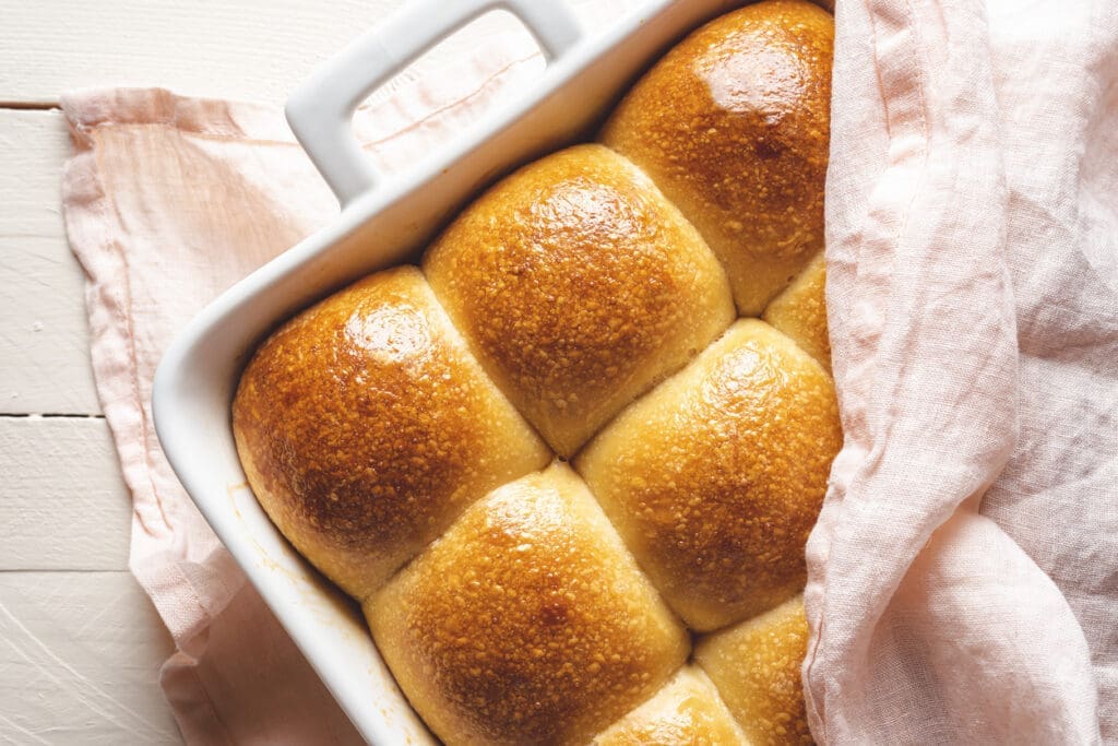 A white baking dish filled with six golden-brown, shiny whole wheat dinner rolls, partially covered by a light pink cloth, sits on a white wooden surface—perfect for holiday baking.