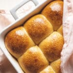 A white baking dish filled with six golden-brown, shiny whole wheat dinner rolls, partially covered by a light pink cloth, sits on a white wooden surface—perfect for holiday baking.