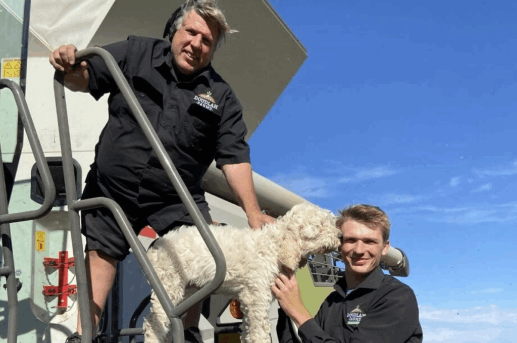 Two men in black shirts stand by farm equipment under a blue sky. As they smile, one shares a personal story about how they began to farm organically, holding a white dog that is being petted by the other man on the stairs.