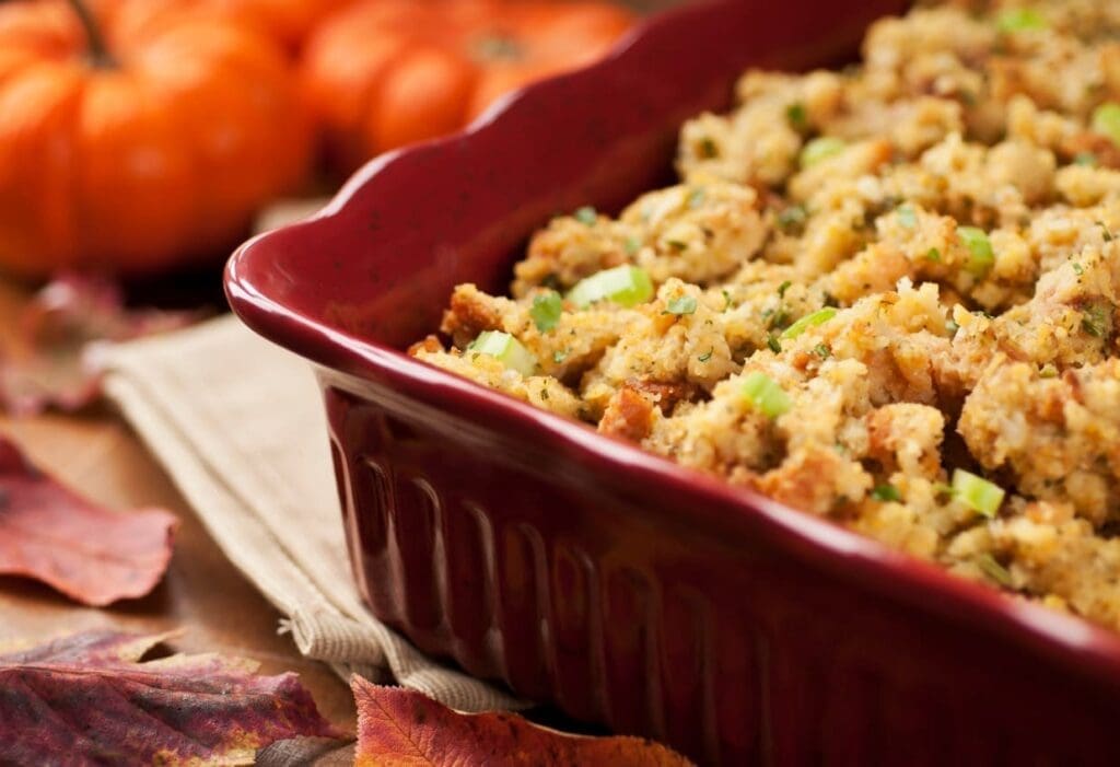 A close-up of a red casserole dish filled with cornbread stuffing, garnished with chopped celery and herbs. Autumn leaves and small pumpkins are blurred in the background, creating a fall-themed setting.