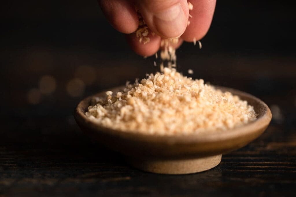 A hand sprinkles a small amount of beige, flaky seasoning into a shallow wooden dish filled with the same seasoning, against a dark, blurred background.