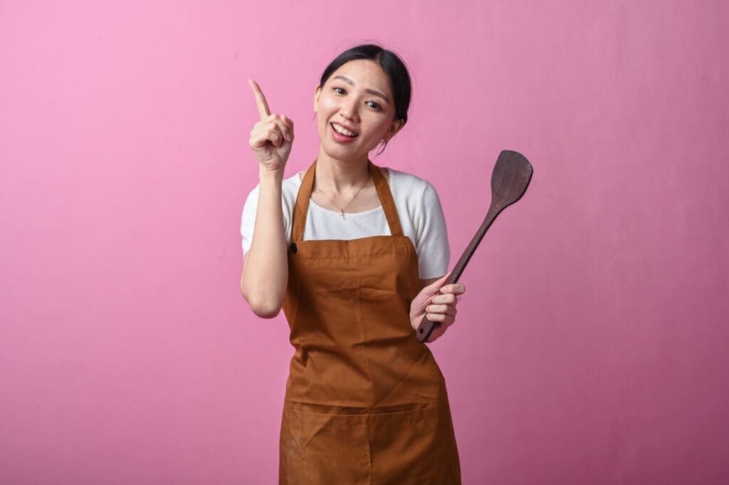A woman wearing a brown apron and white shirt holds a wooden spatula and points upward with her finger, smiling, against a pink background.