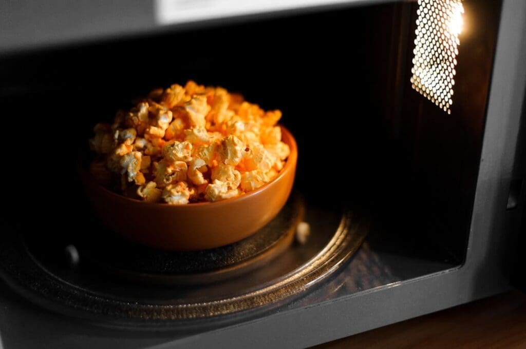 A bowl filled with popcorn sits inside a microwave, with the interior light on and the door partially open.