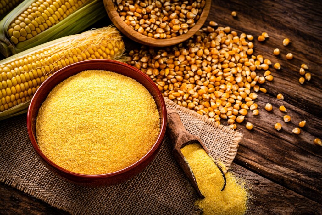 A bowl of cornmeal, a wooden scoop with cornmeal breadcrumbs, corn kernels, and ears of corn arranged on a rustic wooden table with a burlap cloth.