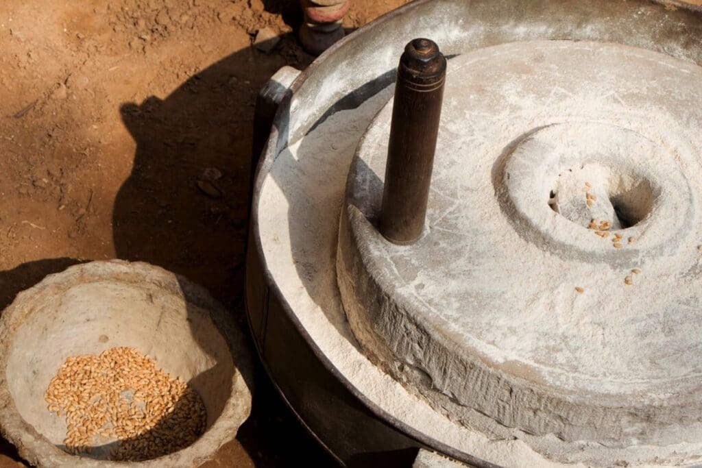 A traditional stone hand mill with grains scattered on its surface, next to a stone bowl filled with whole wheat grains, placed on a dirt ground.
