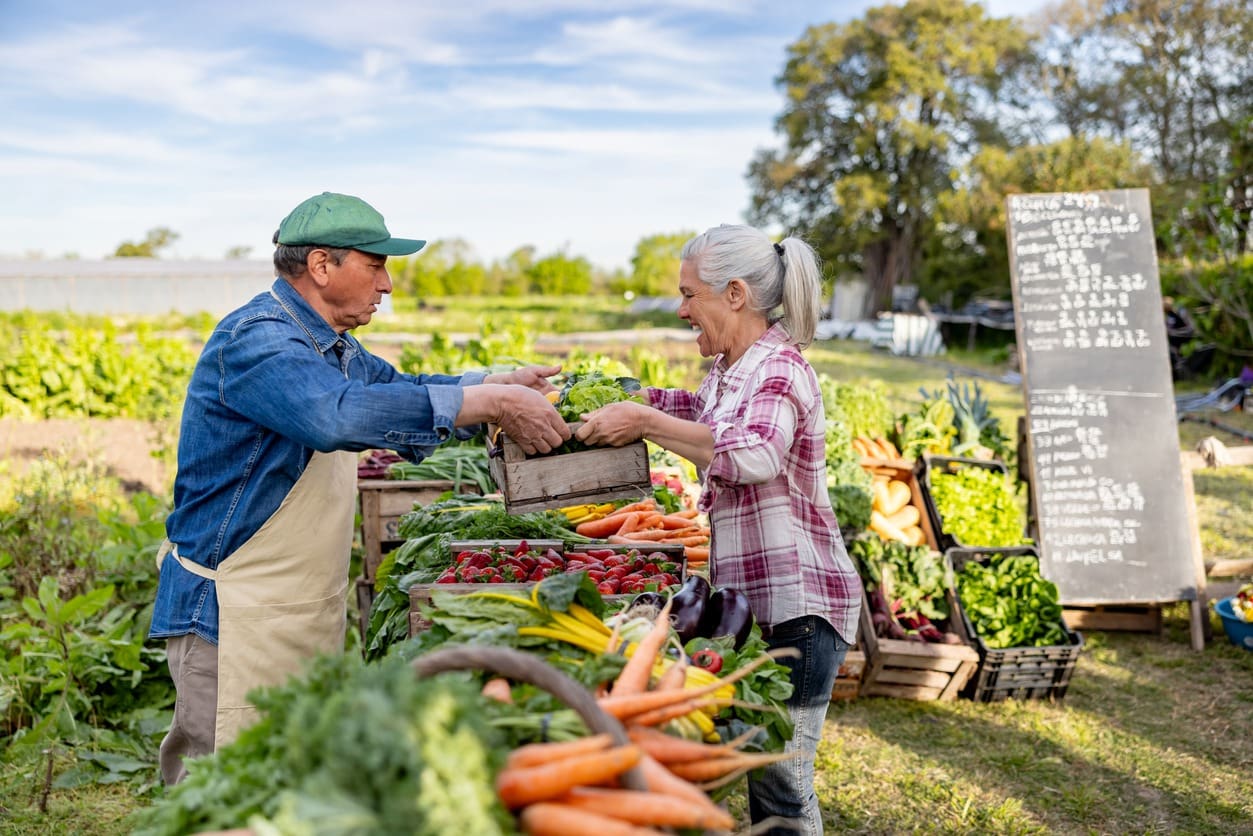 An older man and woman smiling and exchanging a box of fresh vegetables at an outdoor farmers market, surrounded by produce stands and greenery, with a chalkboard menu in the background.