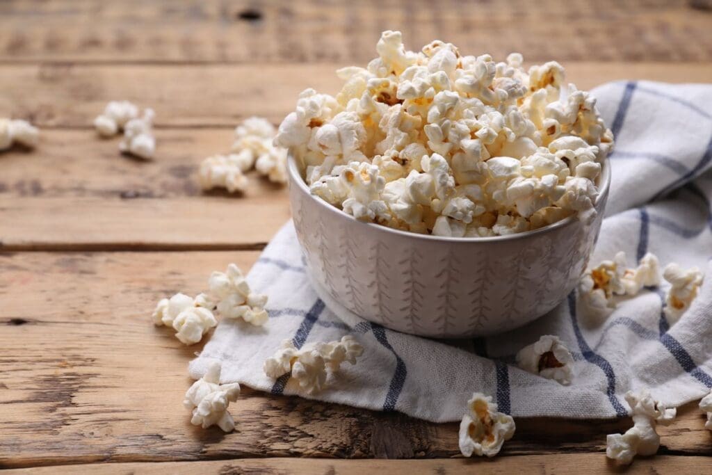A white bowl filled with popcorn sits on a white and blue checkered cloth, with some popcorn scattered around on a rustic wooden table.