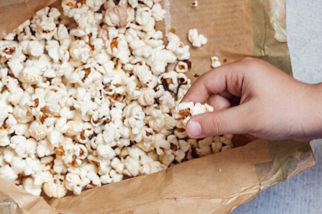 A hand is reaching into a brown paper bag filled with popped popcorn, grabbing a piece from the bunch.