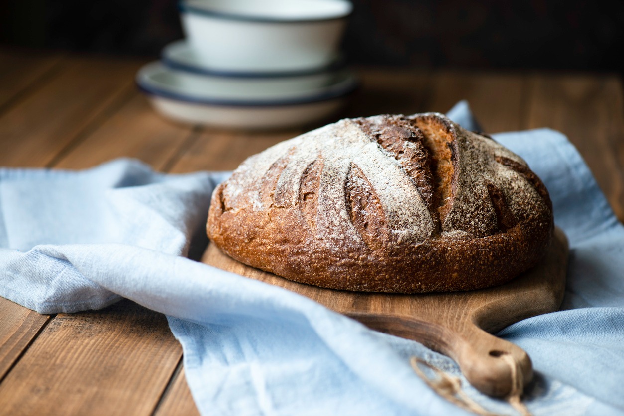 A round loaf of rustic bread made with organic flour sits on a wooden board atop a light blue cloth, with stacked white dishes in the blurred background.
