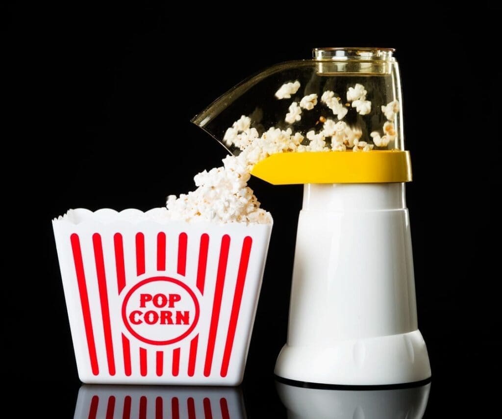 A white popcorn machine pops air-popped popcorn into a red and white striped container labeled Pop Corn against a black background.