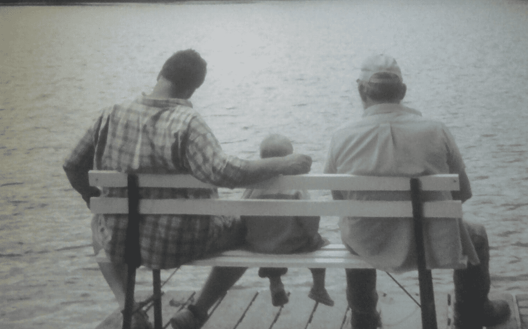 Three people, likely from different generations, sit on a bench facing a body of water. The adult on the left has an arm around a child, while an older person sits on the right. All are viewed from behind.