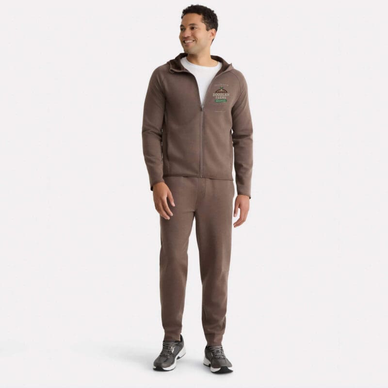 A man smiles against a plain white background, wearing the Premium Hooded Jacket in brown with matching pants, a white t-shirt underneath, and gray sneakers.