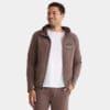 A man smiles wearing a brown Premium Hooded Jacket with the Doubrah Farms logo on the left chest, paired with matching pants and a white T-shirt underneath. One hand is in his pocket.