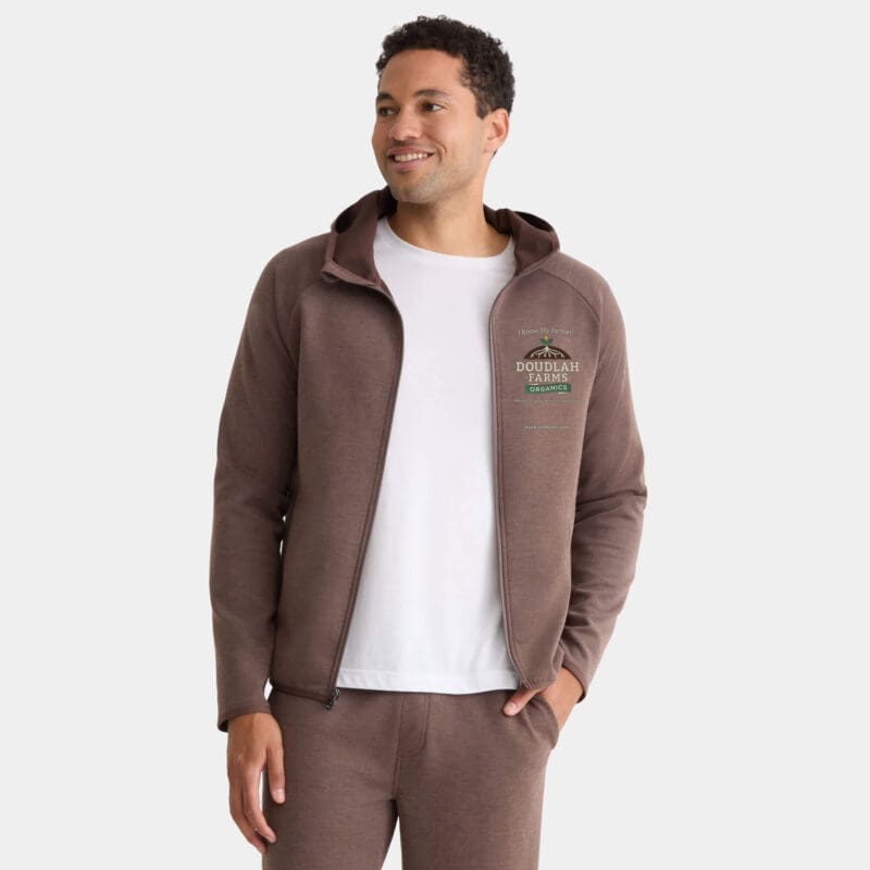 A man smiles wearing a brown Premium Hooded Jacket with the Doubrah Farms logo on the left chest, paired with matching pants and a white T-shirt underneath. One hand is in his pocket.