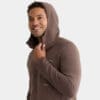 A smiling person wears the Premium Hooded Jacket in brown, holding the hood's edge with one hand and looking off to the side against a plain light background.