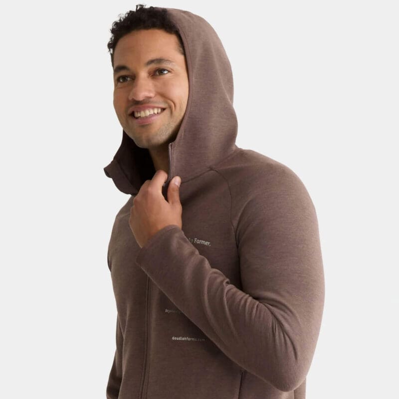 A smiling person wears the Premium Hooded Jacket in brown, holding the hood's edge with one hand and looking off to the side against a plain light background.