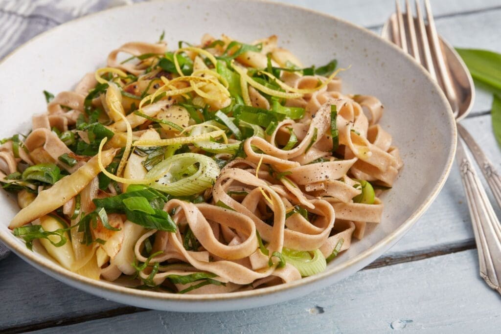 A bowl of spelt fettuccine pasta topped with sautéed vegetables, fresh greens, asparagus, and lemon zest, garnished with black pepper, sits on a blue wooden table next to a fork and knife.