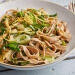 A bowl of spelt fettuccine pasta topped with sautéed vegetables, fresh greens, asparagus, and lemon zest, garnished with black pepper, sits on a blue wooden table next to a fork and knife.