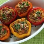 Five stuffed bell peppers, red and orange, filled with a rice, pinto bean, and vegetable mixture, topped with melted cheese and chopped herbs, sit in a white oval dish on a green placemat—a perfect spring recipe.