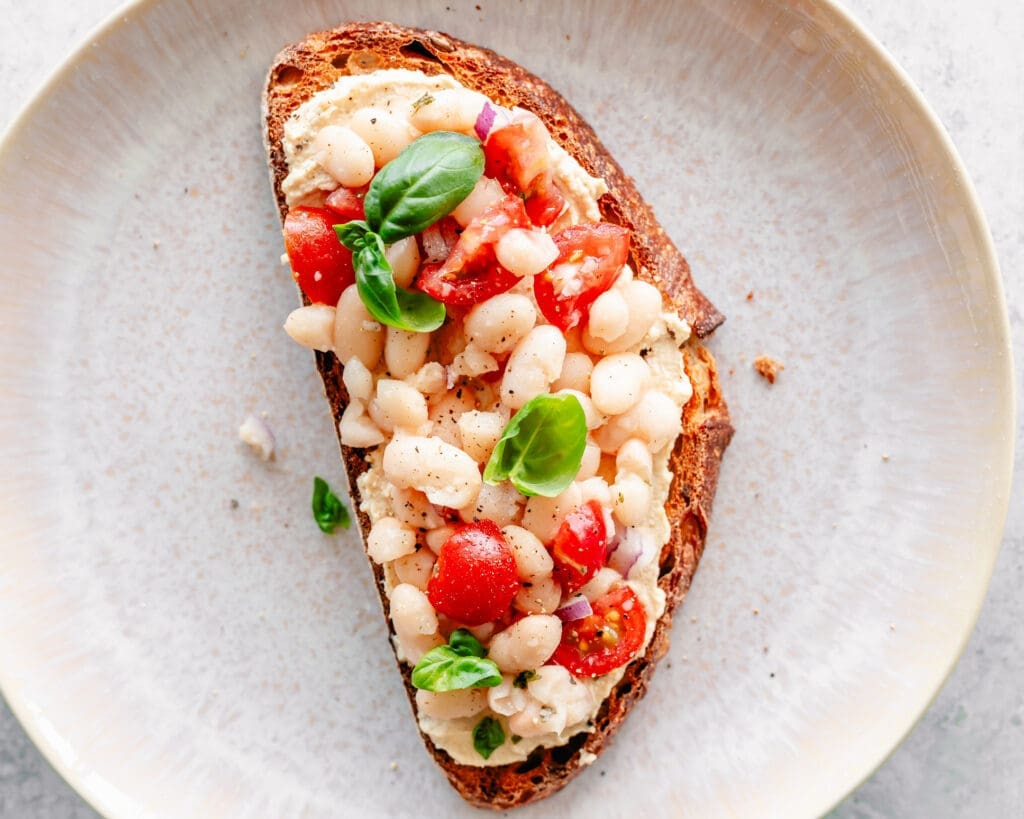 A slice of toasted bread topped with organic navy beans, diced tomatoes, red onion, fresh basil leaves, and a spread, served on a beige plate.