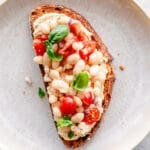 A slice of toasted bread topped with organic navy beans, diced tomatoes, red onion, fresh basil leaves, and a spread, served on a beige plate.