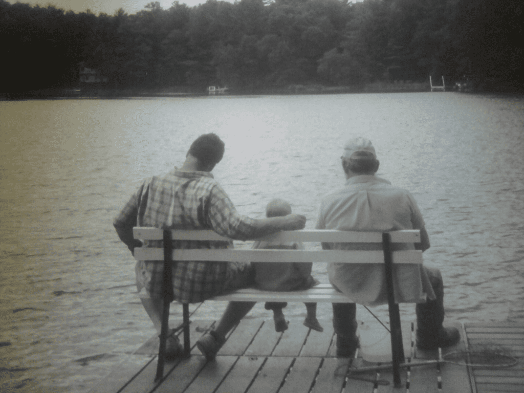 Three people—an adult, a child, and an older adult—sit on a bench at the edge of a dock, facing a calm lake with trees in the background. The photo is in black and white.