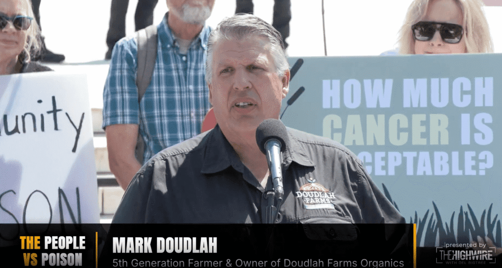 A man speaks at a podium with a microphone. Behind him, people hold protest signs, one asking “HOW MUCH CANCER IS ACCEPTABLE?” Among the crowd, concerns about mantle cell lymphoma are apparent; onscreen text identifies the speaker as Mark Doudlah.