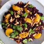 A bowl of black rice salad topped with mandarin orange slices, cilantro leaves, edamame, and slivered almonds. Fresh ginger, cilantro, black rice, and almonds are scattered around the bowl with a side of ginger soy dressing.