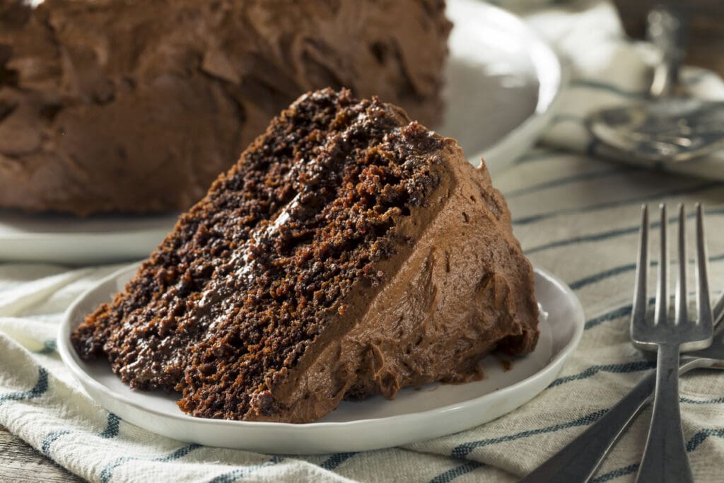 A close-up of a slice of rich, moist chocolate cake with thick chocolate frosting on a small white plate, set on a striped cloth napkin with a fork beside it.