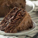 A close-up of a slice of rich, moist chocolate cake with thick chocolate frosting on a small white plate, set on a striped cloth napkin with a fork beside it.