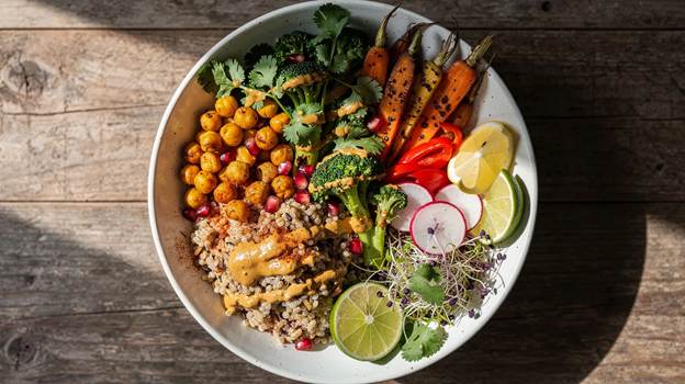 A colorful bowl of quinoa topped with roasted chickpeas, carrots, broccoli, red bell peppers, radishes, lime slices, microgreens, cilantro, pomegranate seeds, and a drizzle of creamy dressing on a wooden table.