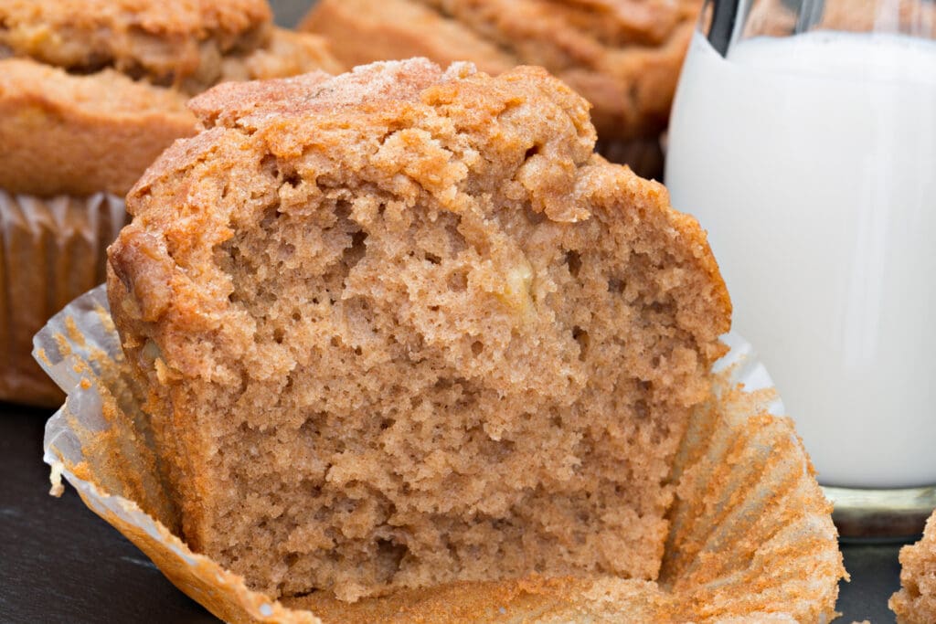 A close-up of a cinnamon muffin with a soft, fluffy texture, partially unwrapped from its paper liner, sits next to a glass of milk. Other muffins are visible in the background.