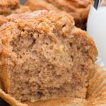 A close-up of a cinnamon muffin with a soft, fluffy texture, partially unwrapped from its paper liner, sits next to a glass of milk. Other muffins are visible in the background.