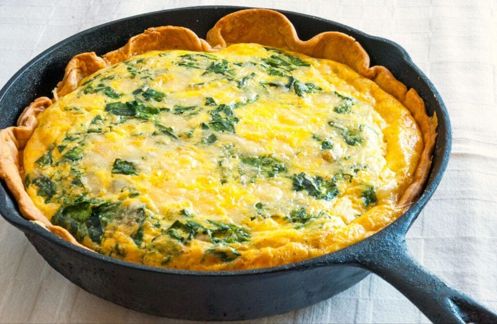 A baked spinach and cheese quiche with a golden crust sits in a black cast iron skillet on a white tablecloth.