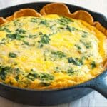 A baked spinach and cheese quiche with a golden crust sits in a black cast iron skillet on a white tablecloth.
