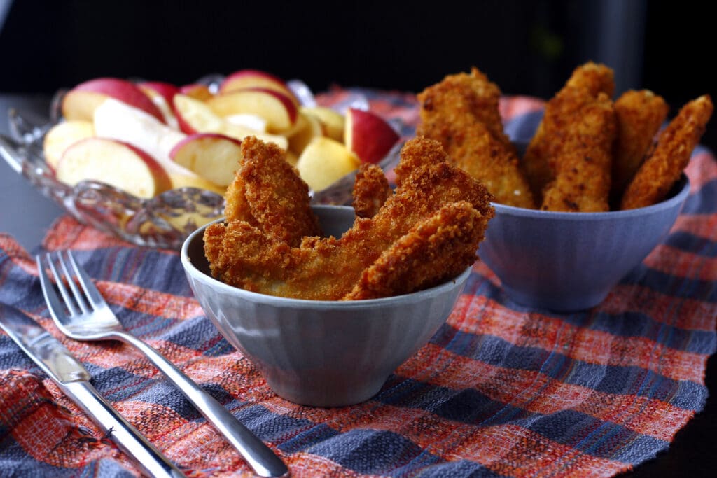 Two bowls filled with crispy breaded chicken tenders sit on a plaid cloth, with a plate of sliced apples and a fork and knife nearby. The background is softly lit.
